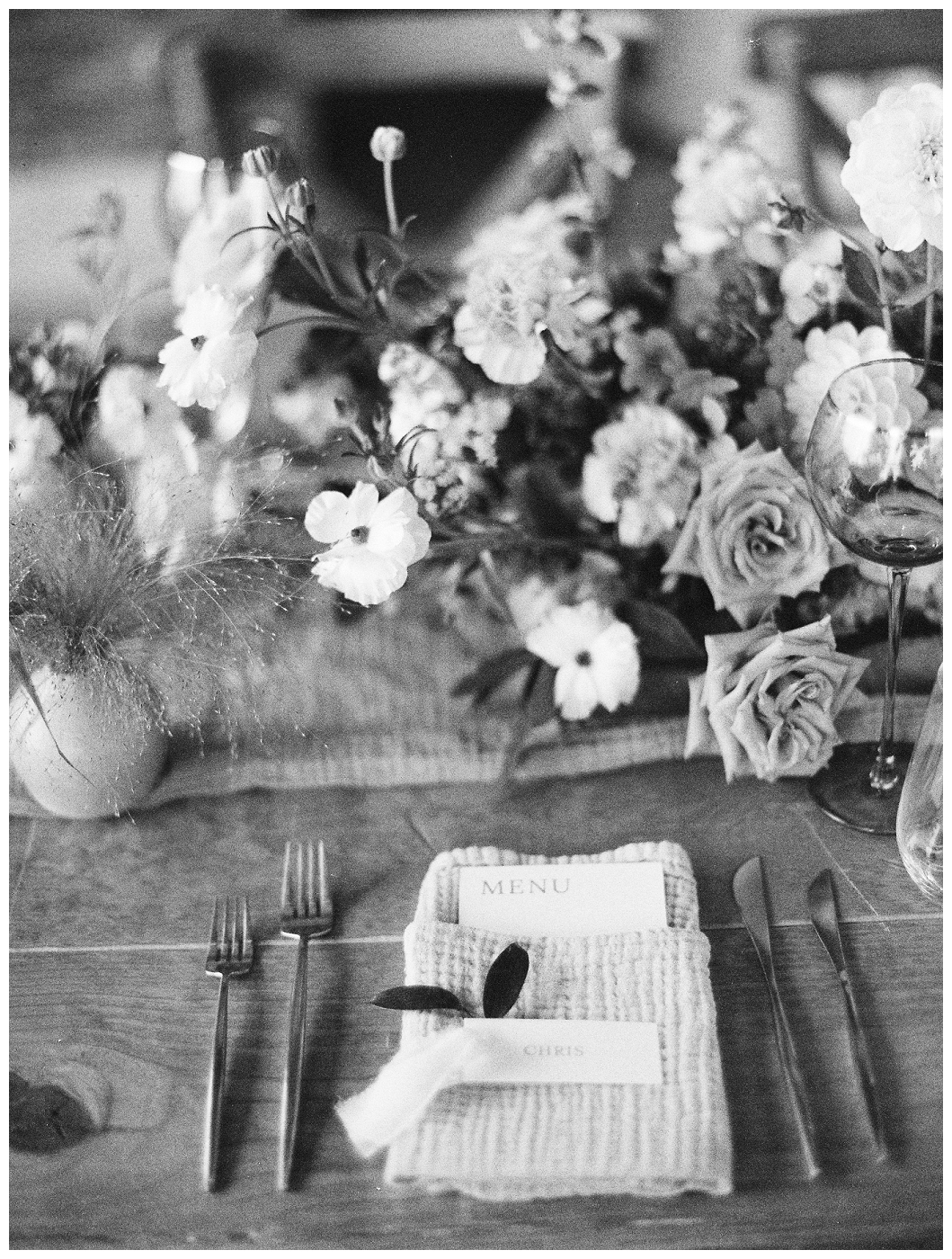 black and white photo of reception table place setting with centerpiece and budvase, del mar surf station, farmhouse table, budvases ,outdoor reception, medium sized centerpieces, romantic weddings, daisies, toffee roses, berries, cream flowers, white flowers, hydrangeas, majolica roses, explosion grass, outdoor wedding, covid wedding, san diego wedding florist, southern california elopement florist, socal florist, best wedding florist san diego ca, best wedding florist southern california, los angeles wedding florist, top rated wedding florist in california, del mar san diego, blush tapered candles, blush table runner, budvases