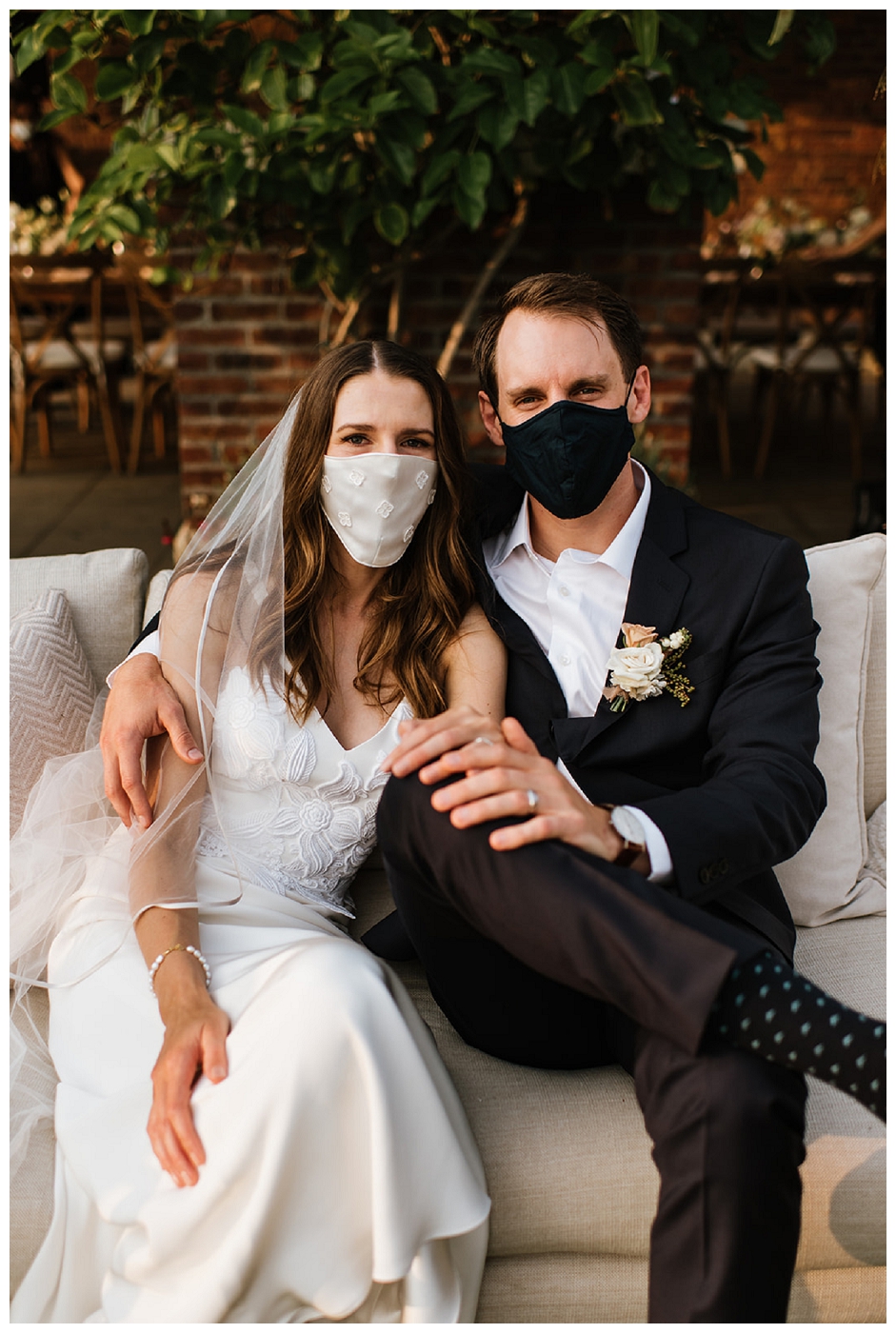 bride and groom posing together in masks cuddle up on a couch, groom wearing a boutonniere, del mar surf station, farmhouse table, budvases ,outdoor reception, medium sized centerpieces, romantic weddings, daisies, toffee roses, berries, cream flowers, white flowers, hydrangeas, majolica roses, explosion grass, outdoor wedding, covid wedding, san diego wedding florist, southern california elopement florist, socal florist, best wedding florist san diego ca, best wedding florist southern california, los angeles wedding florist, top rated wedding florist in california, del mar san diego, blush tapered candles, blush table runner, budvases