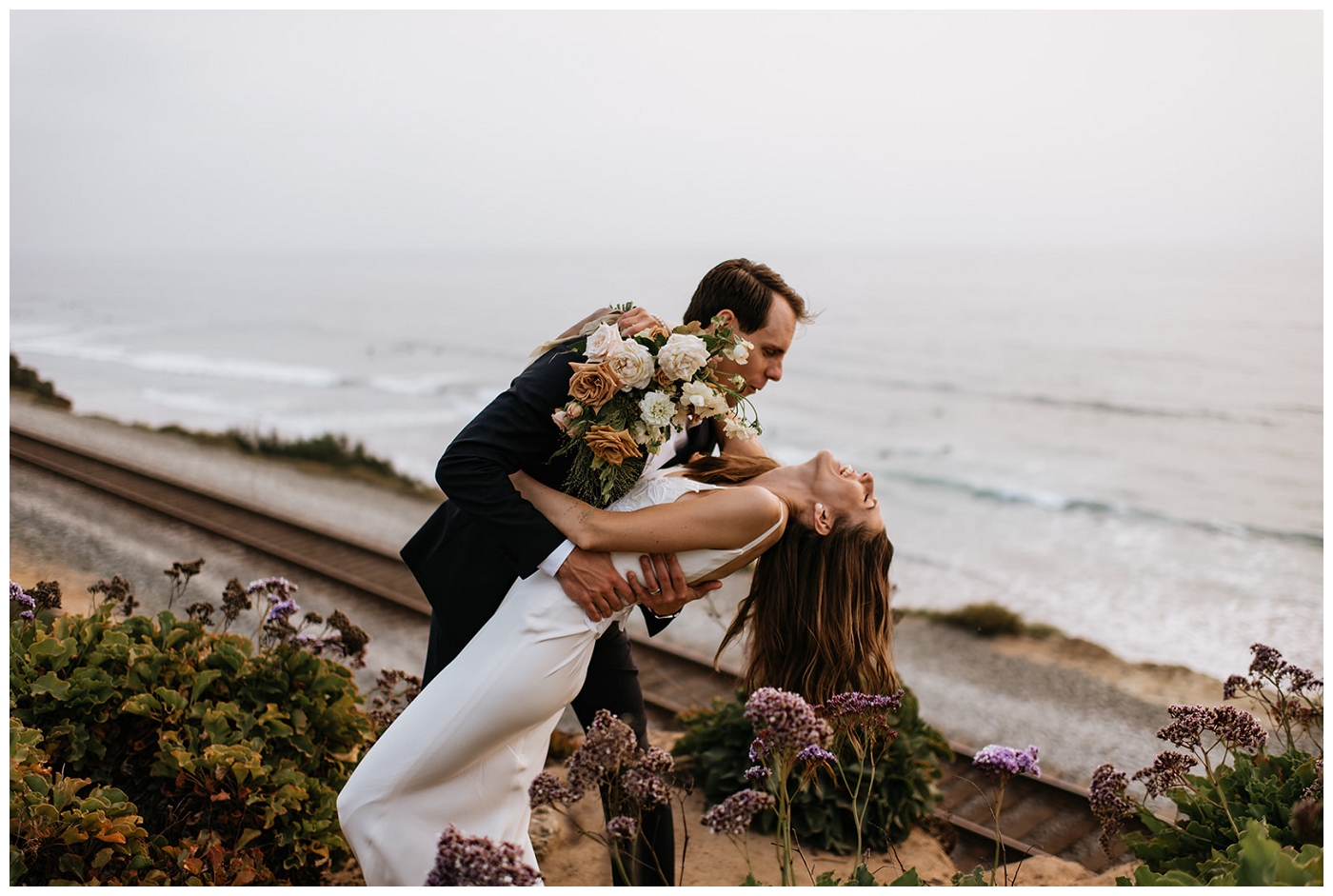 groom dipping his bride in front of beach view while she holds her bouquet, del mar surf station, farmhouse table, budvases ,outdoor reception, medium sized centerpieces, romantic weddings, daisies, toffee roses, berries, cream flowers, white flowers, hydrangeas, majolica roses, explosion grass, outdoor wedding, covid wedding, san diego wedding florist, southern california elopement florist, socal florist, best wedding florist san diego ca, best wedding florist southern california, los angeles wedding florist, top rated wedding florist in california, del mar san diego, blush tapered candles, blush table runner, budvases