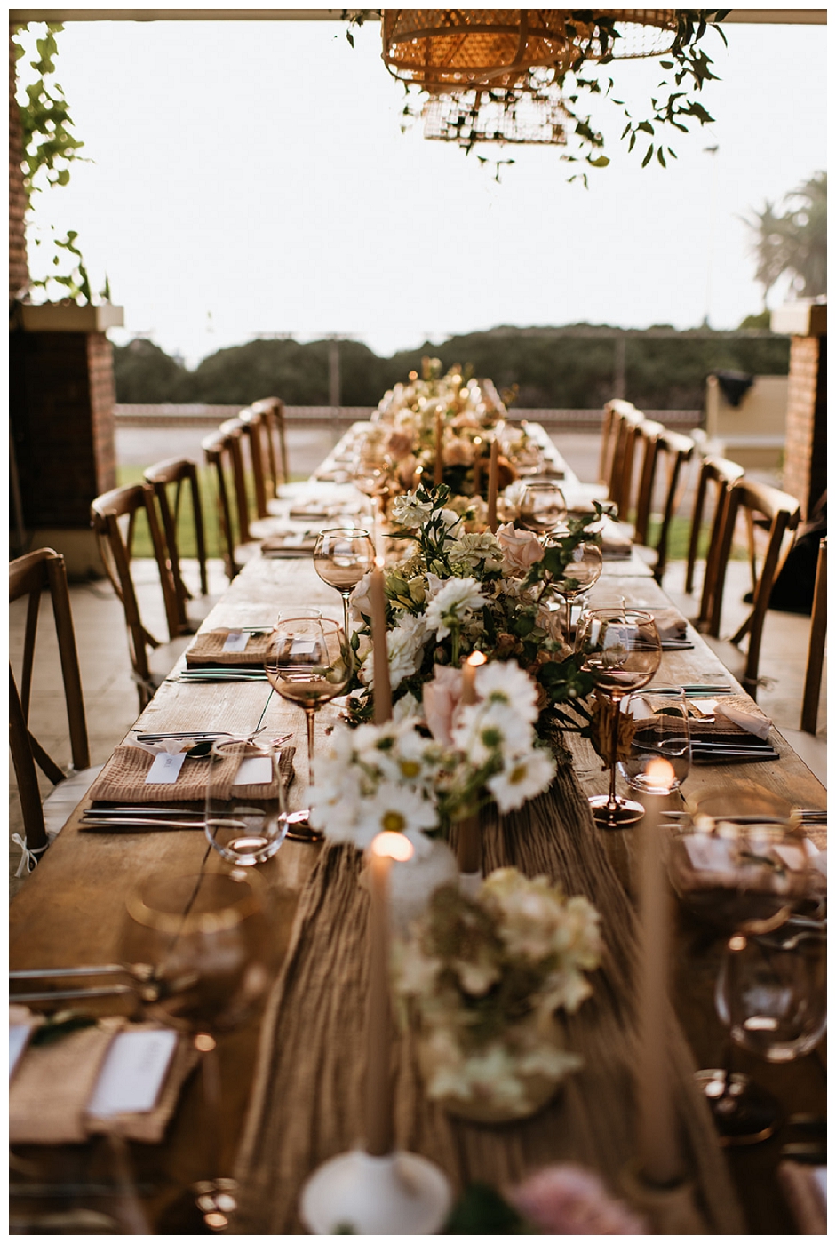 long shot of farmhouse table wedding reception topped with blush runner and floral centerpieces surrounded by tapered candles, del mar surf station, farmhouse table, budvases ,outdoor reception, medium sized centerpieces, romantic weddings, daisies, toffee roses, berries, cream flowers, white flowers, hydrangeas, majolica roses, explosion grass, outdoor wedding, covid wedding, san diego wedding florist, southern california elopement florist, socal florist, best wedding florist san diego ca, best wedding florist southern california, los angeles wedding florist, top rated wedding florist in california, del mar san diego, blush tapered candles, blush table runner, budvases